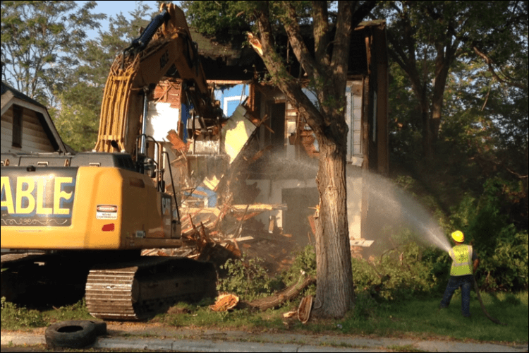 A front-end loader is used to demolish an abandoned residential dwelling in Detroit, where asbestos may be present, while a man in safety gear sprays water. 