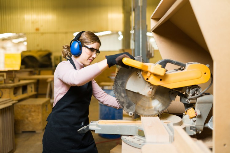 A woman wears hearing protection and safety glasses while using a bench saw to cut a board.