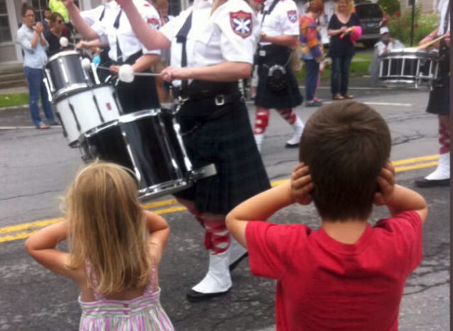 Two children cover their ears as drummers in a parade march by.